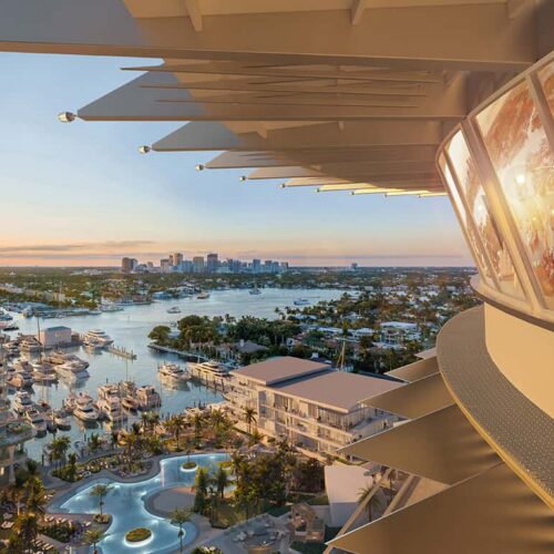 Aerial view of a waterfront resort with a marina of boats, palm trees, and a city skyline at sunset; a curved glass-walled restaurant extends from the right.