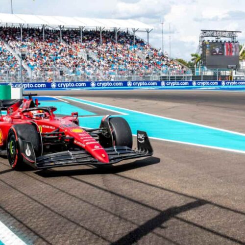 Red Ferrari Formula 1 car speeding along a race track with turquoise run-off and a packed grandstand behind Rolex banners.