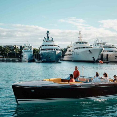 Open-top wooden speedboat carrying five people cruising past a marina with luxury white yachts in the background, under a sunny sky