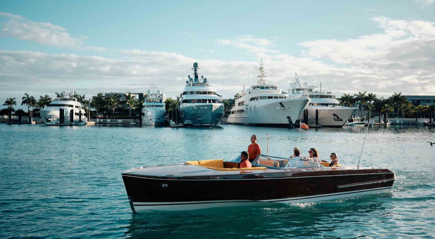 Open-top wooden speedboat carrying five people cruising past a marina with luxury white yachts in the background, under a sunny sky
