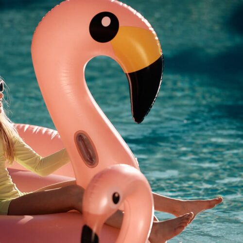 Girl in a pale yellow swimsuit sits on a pink flamingo pool float, wearing white sunglasses and holding a palm leaf.