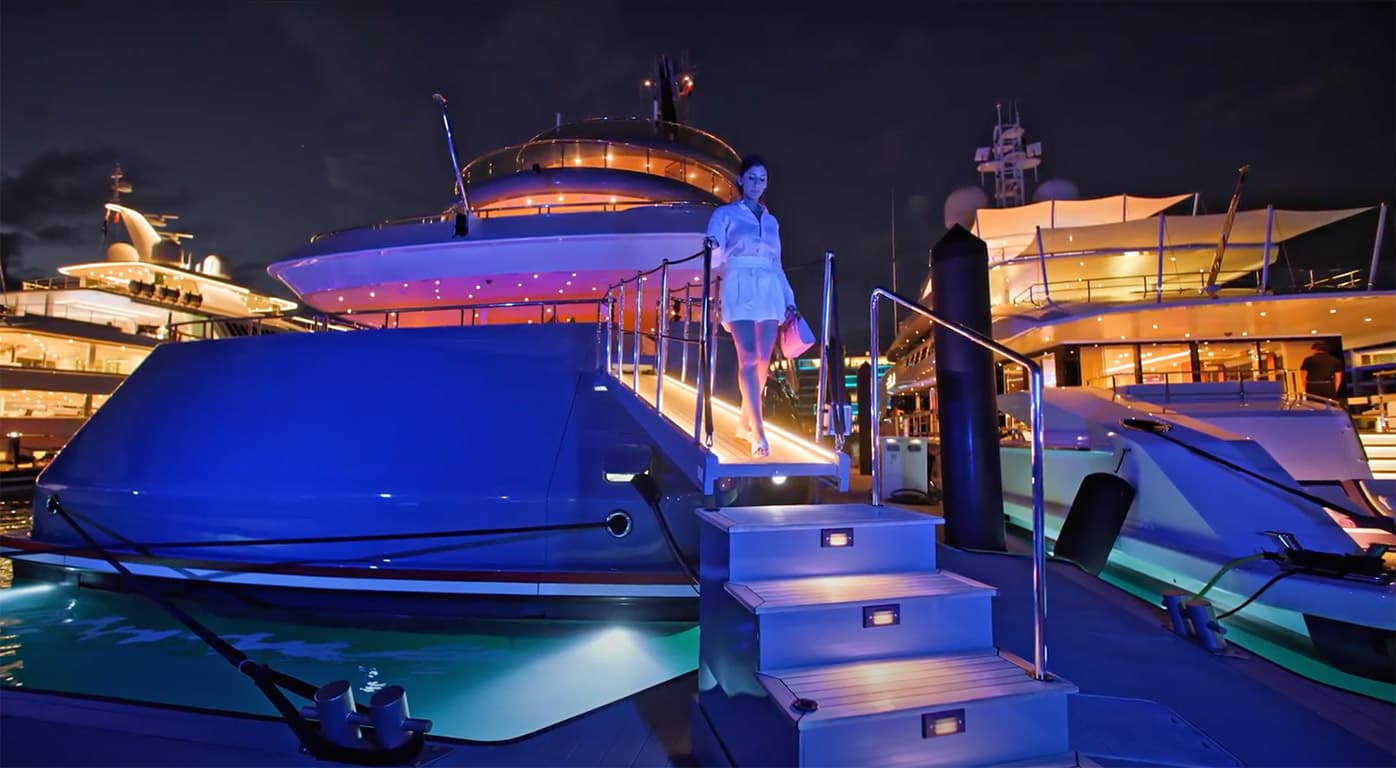 Woman in a light blue dress stepping down a lit staircase onto a luxury yacht at night.