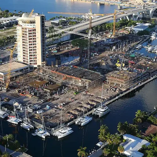 Harbor construction site with cranes and a tall building under development beside a marina of yachts and a bridge in the background