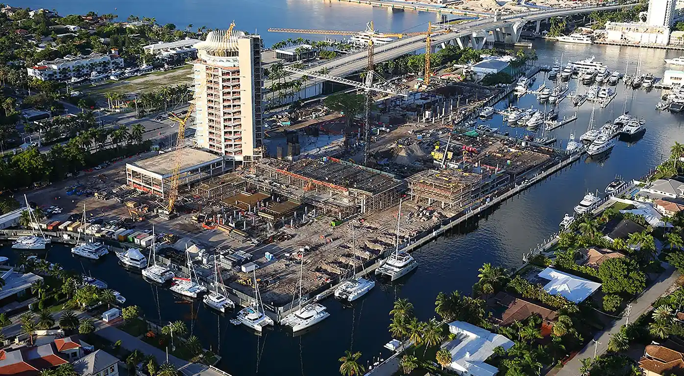 Harbor construction site with cranes and a tall building under development beside a marina of yachts and a bridge in the background