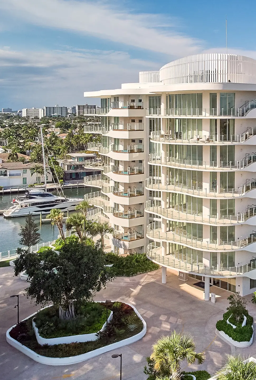 White circular apartment building with glass balconies by a marina; yachts docked nearby and palm trees in the foreground.
