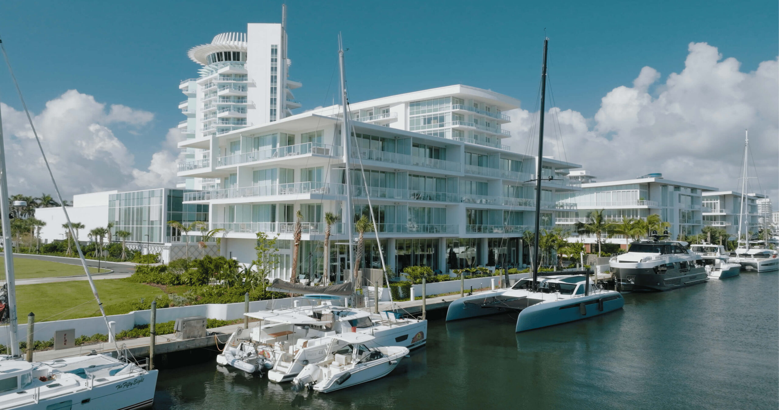 White multi-story waterfront condo buildings with balconies along a marina, yachts docked in the foreground on a sunny day.