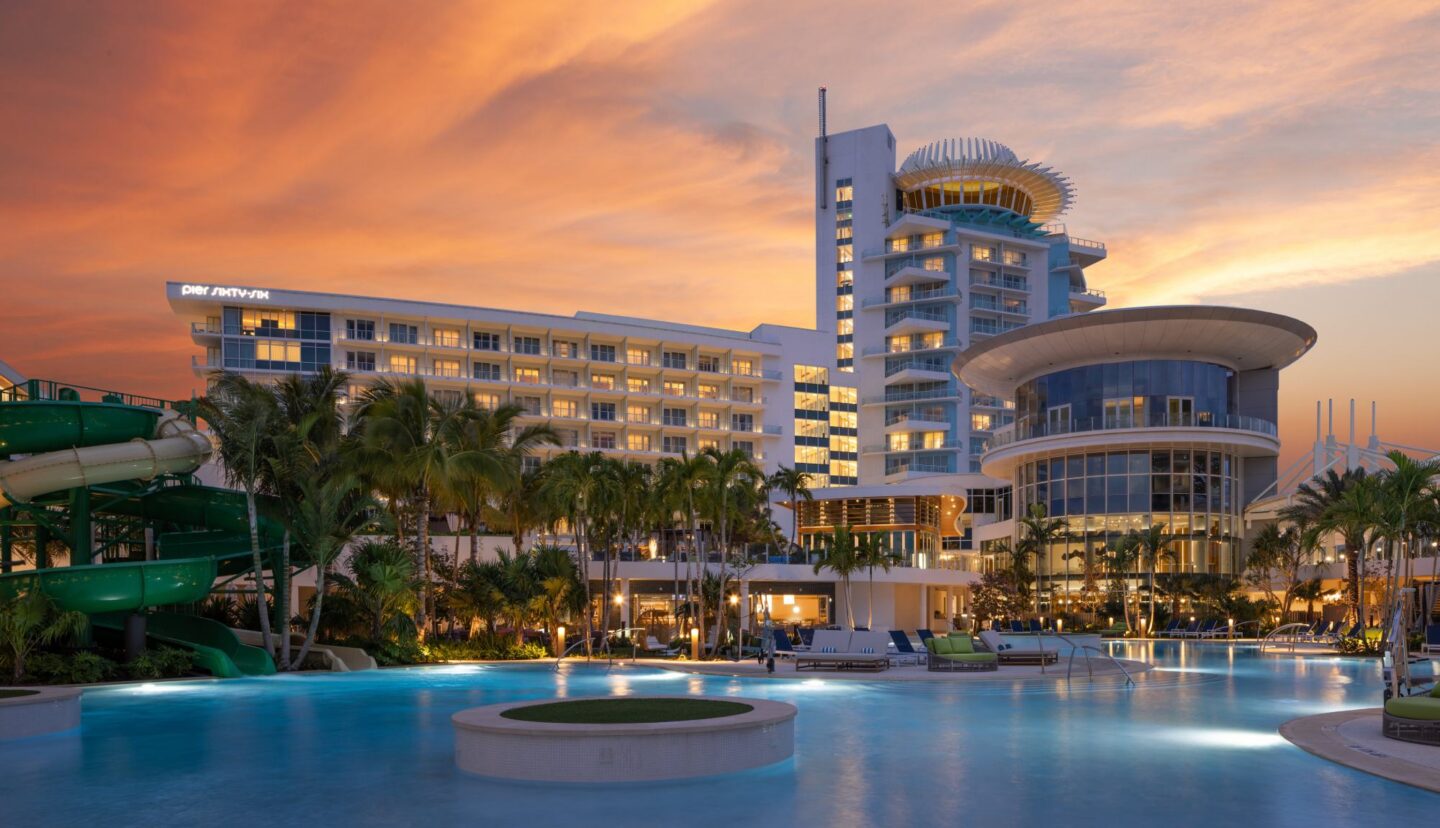 Sunset-lit resort with a large pool, palm trees, and a modern multi-story hotel building.