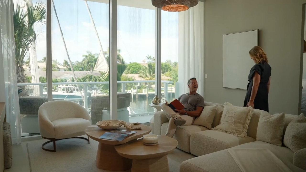 A man sits on a light sofa reading a red notebook while a woman stands nearby in a modern bright living room overlooking a balcony and waterway.