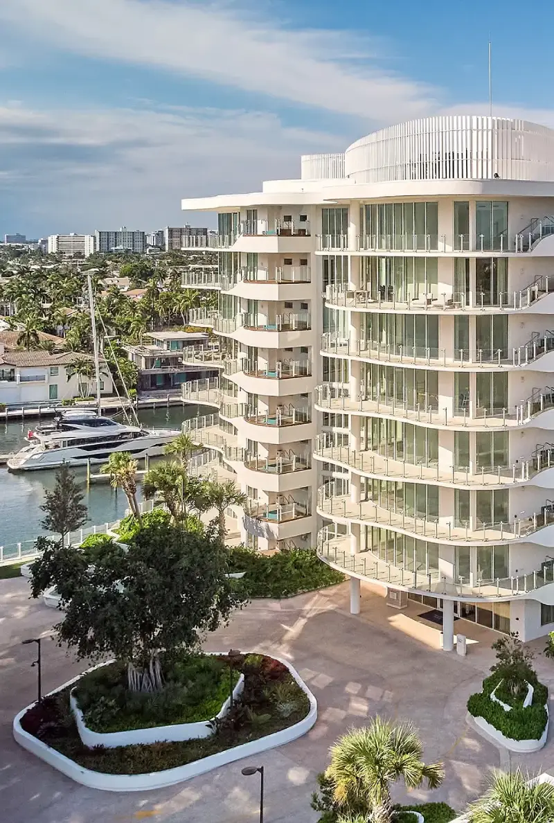 White circular apartment building with glass balconies by a marina; yachts docked nearby and palm trees in the foreground.