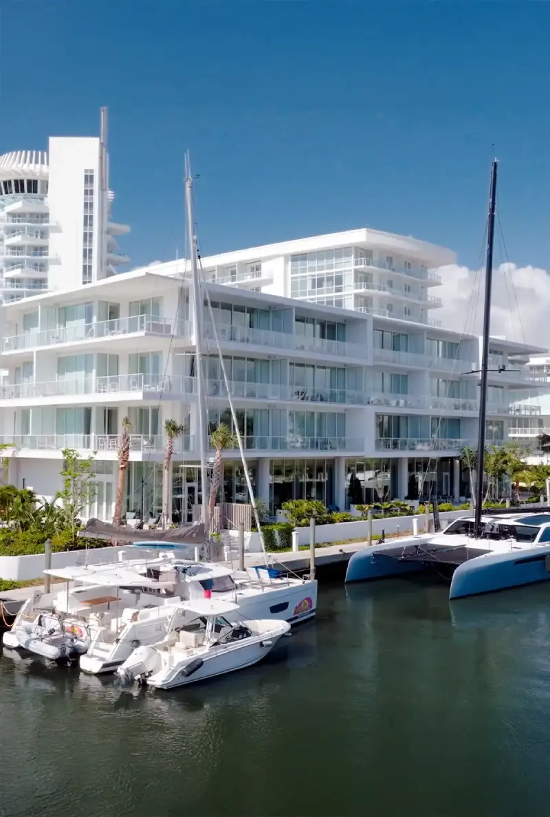 Modern white waterfront apartment building with glass balconies along a marina, with several sailboats docked at the pier beneath a clear blue sky.