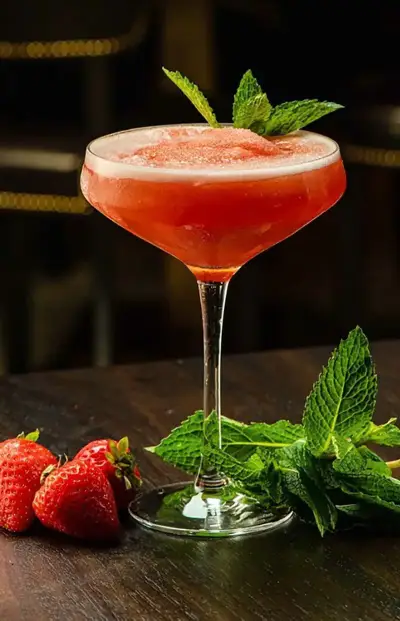 Pink cocktail in a coupe glass with a mint sprig garnish, on a dark bar; fresh strawberries and mint leaves sit on the table nearby.